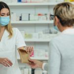 Female pharmacist wearing protective mask and serving a customer patient in a pharmacy and packing drugs in a paper bag