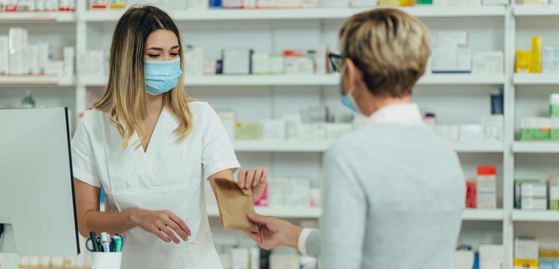 Female pharmacist wearing protective mask and serving a customer patient in a pharmacy and packing drugs in a paper bag